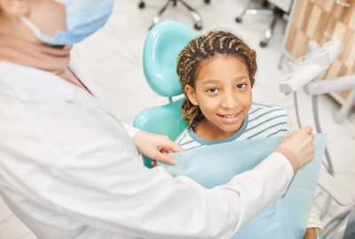 Portrait of little patient smiling at camera sitting on dental chair while dentist preparing her to medical exam